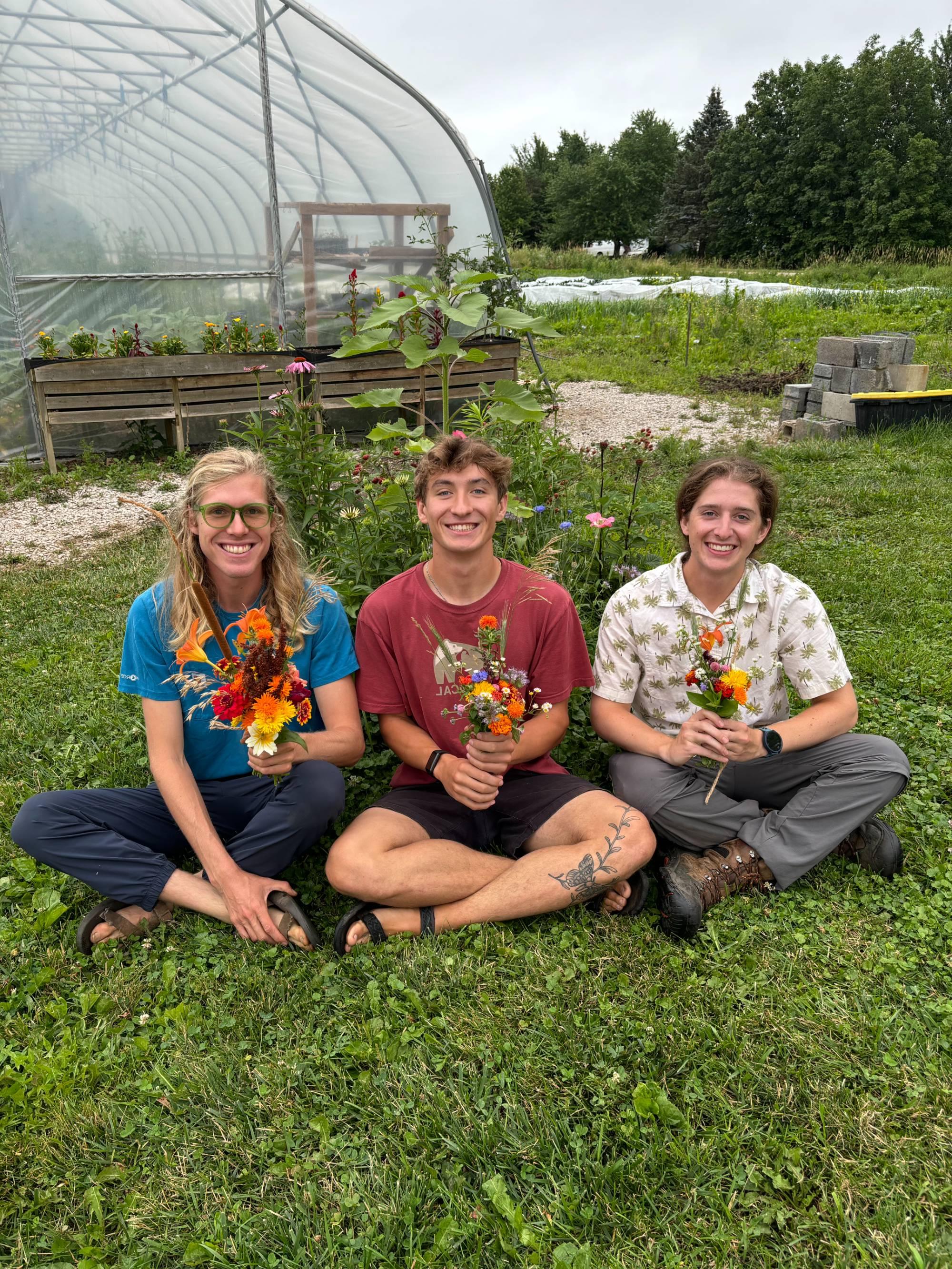 Three students sitting on the ground outside each holding a bouquet of flowers